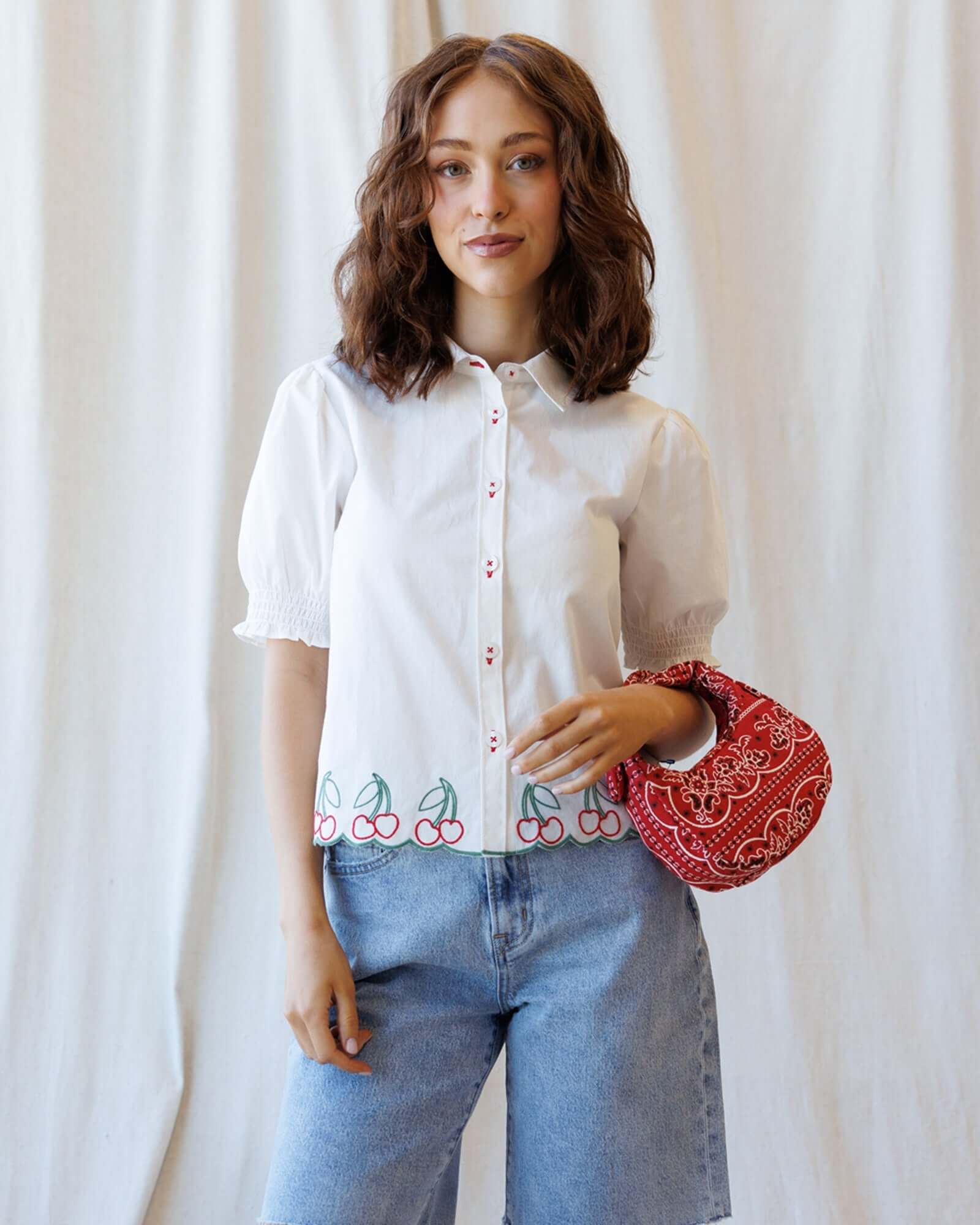Cherry Cute Button Down featuring a cherry embroidered scallop hem and short puff sleeves, styled with a red bag.