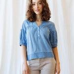 Woman wearing a blue denim shirt and beige pants against a white curtain background