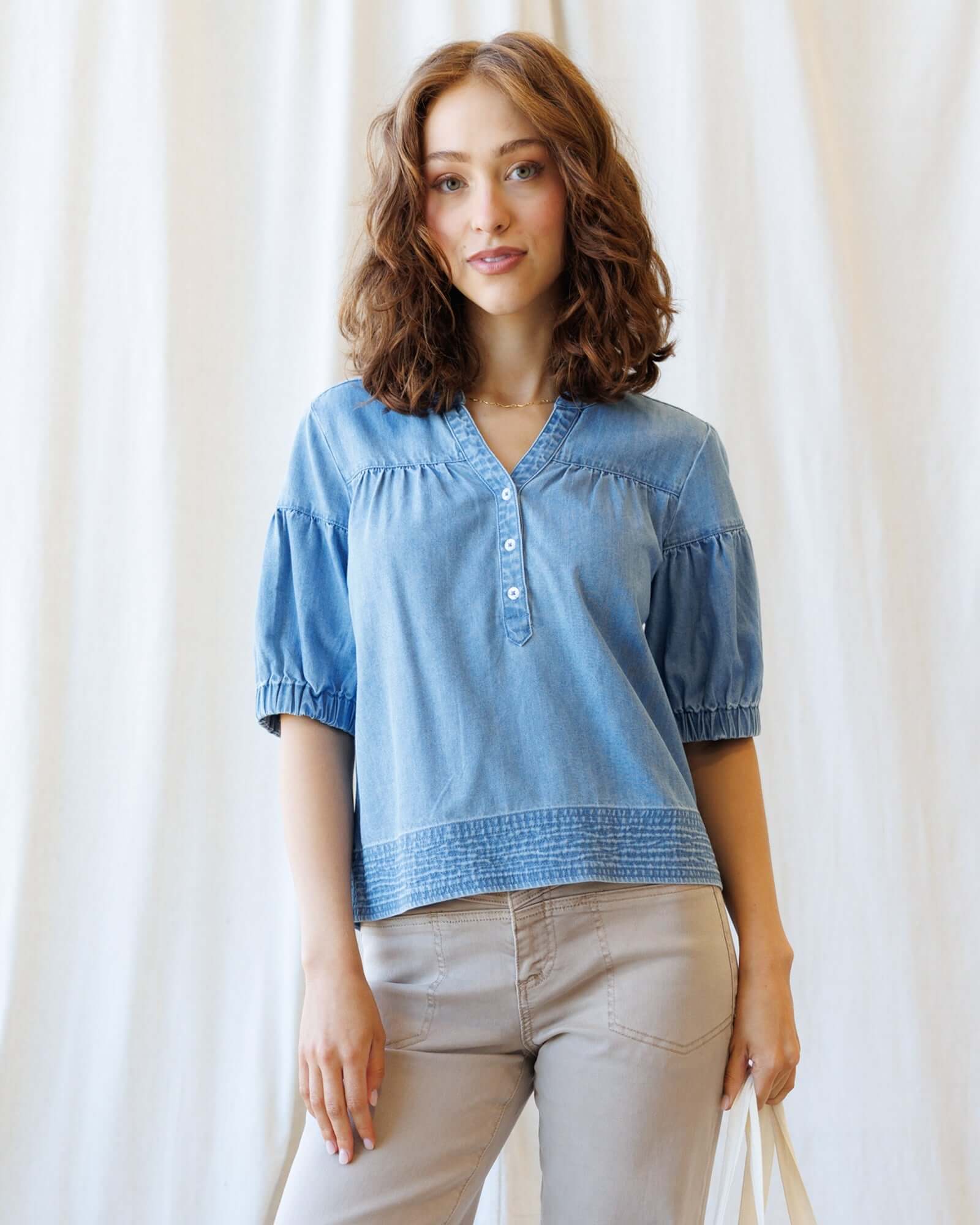 Woman wearing a blue denim shirt and beige pants against a white curtain background