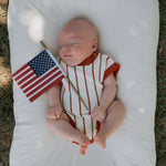 Baby peacefully sleeping in a Picnic Pinstripe | Knit Romper while holding a small American flag on a soft surface.