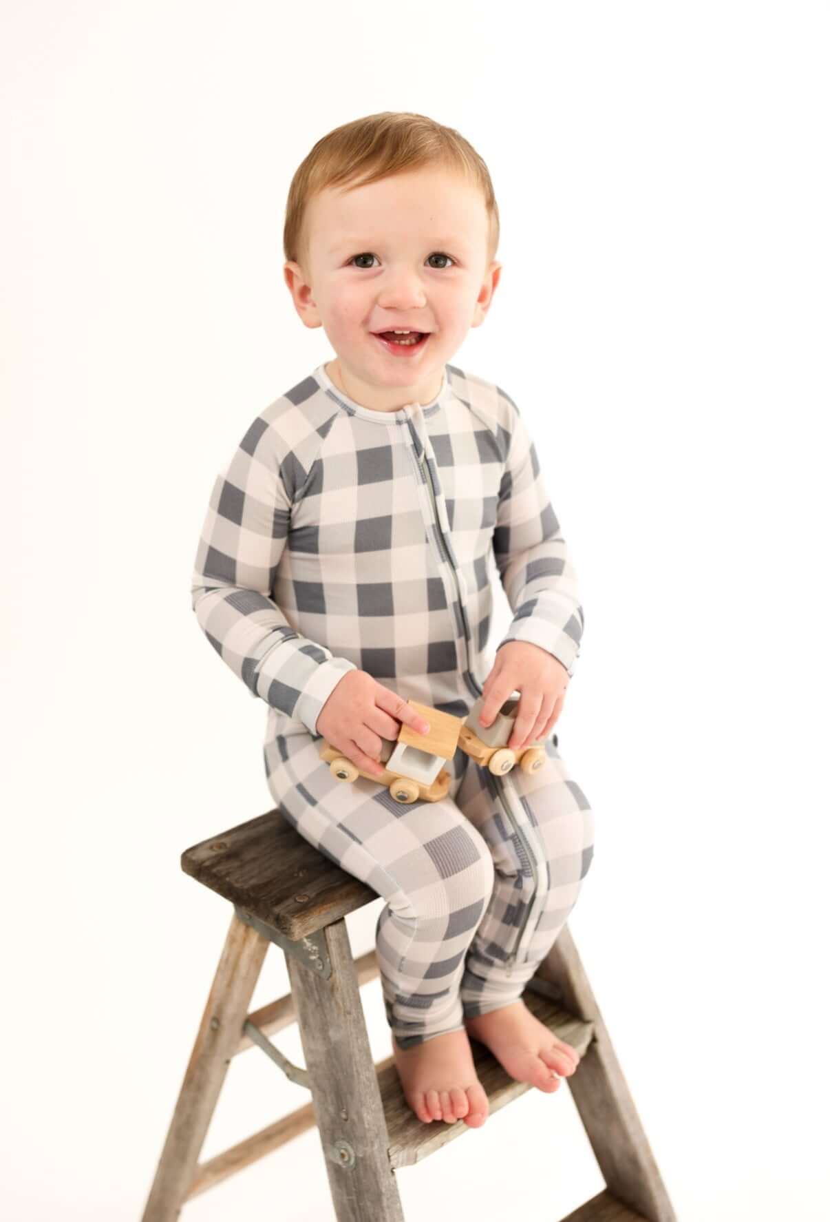 Happy child wearing Slate Gingham | Zip Romper, sitting on a stool holding toy trucks, showcasing adorable design.
