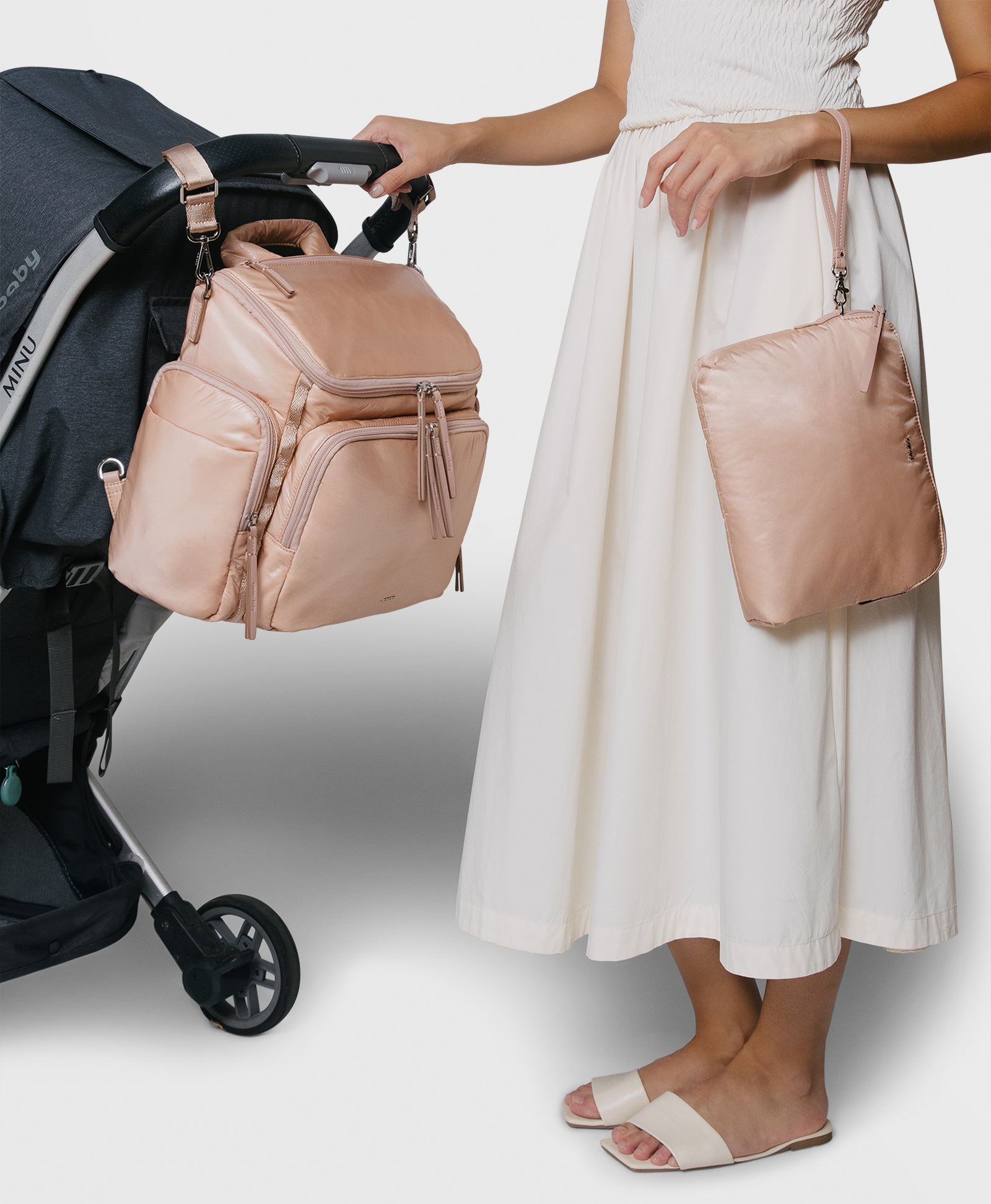 Caraa Baby Bag in blush nylon hanging from stroller, held by a woman in a white skirt, showcasing its design and functionality.