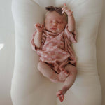 Newborn baby in a pink checkered outfit and headband lying on a white cushion.