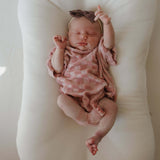 Newborn baby in a pink checkered outfit and headband lying on a white cushion.