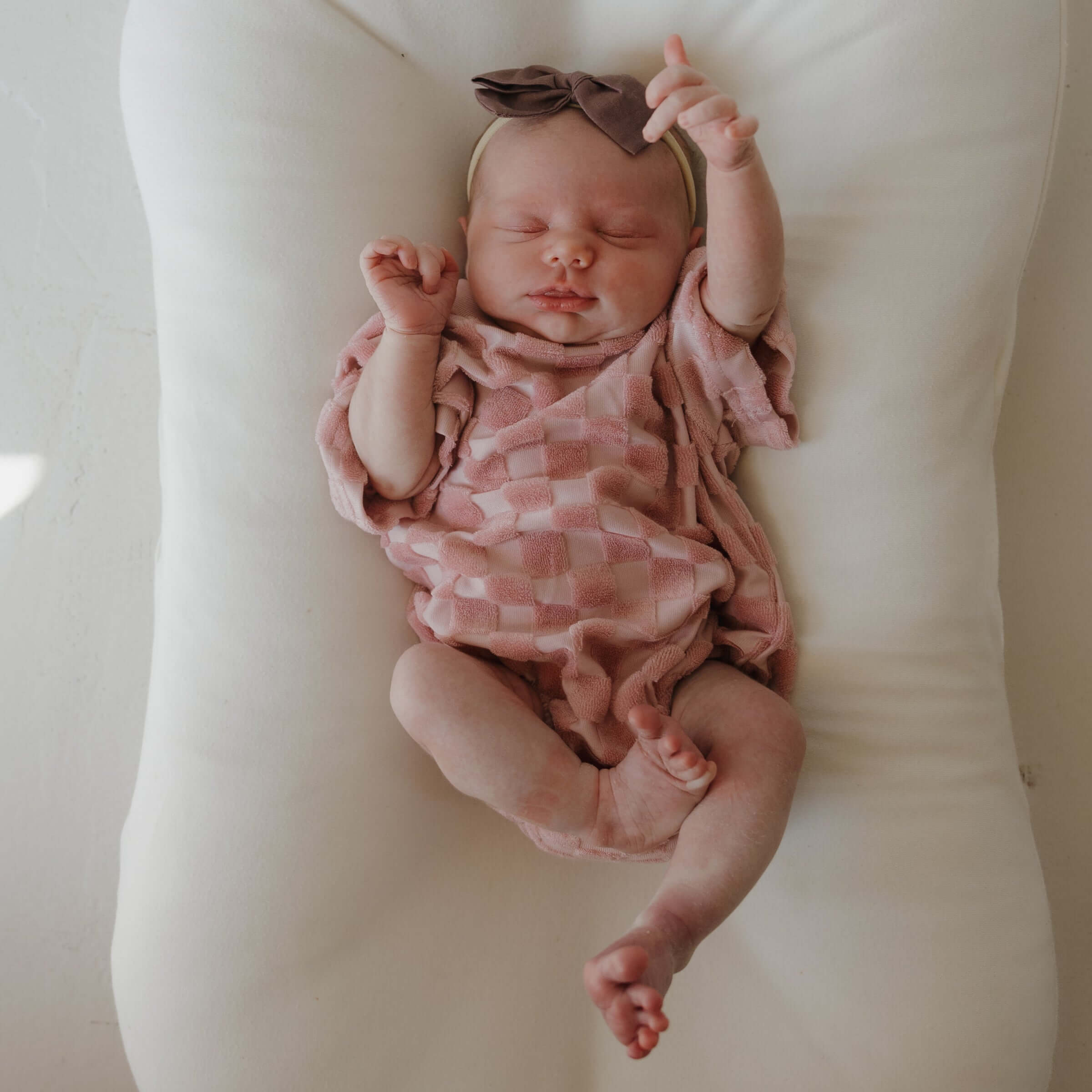 Newborn baby in a pink checkered outfit and headband lying on a white cushion.