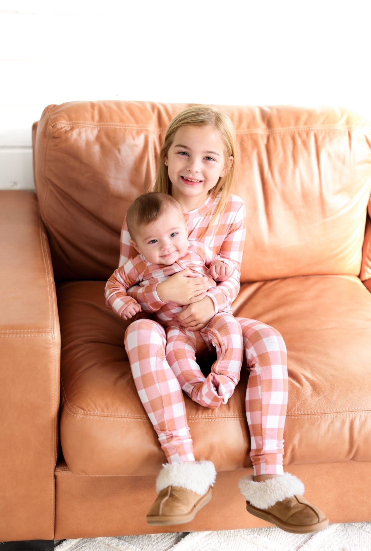 Two children in Coral Gingham zip rompers sitting together on a couch, showcasing comfort and style.