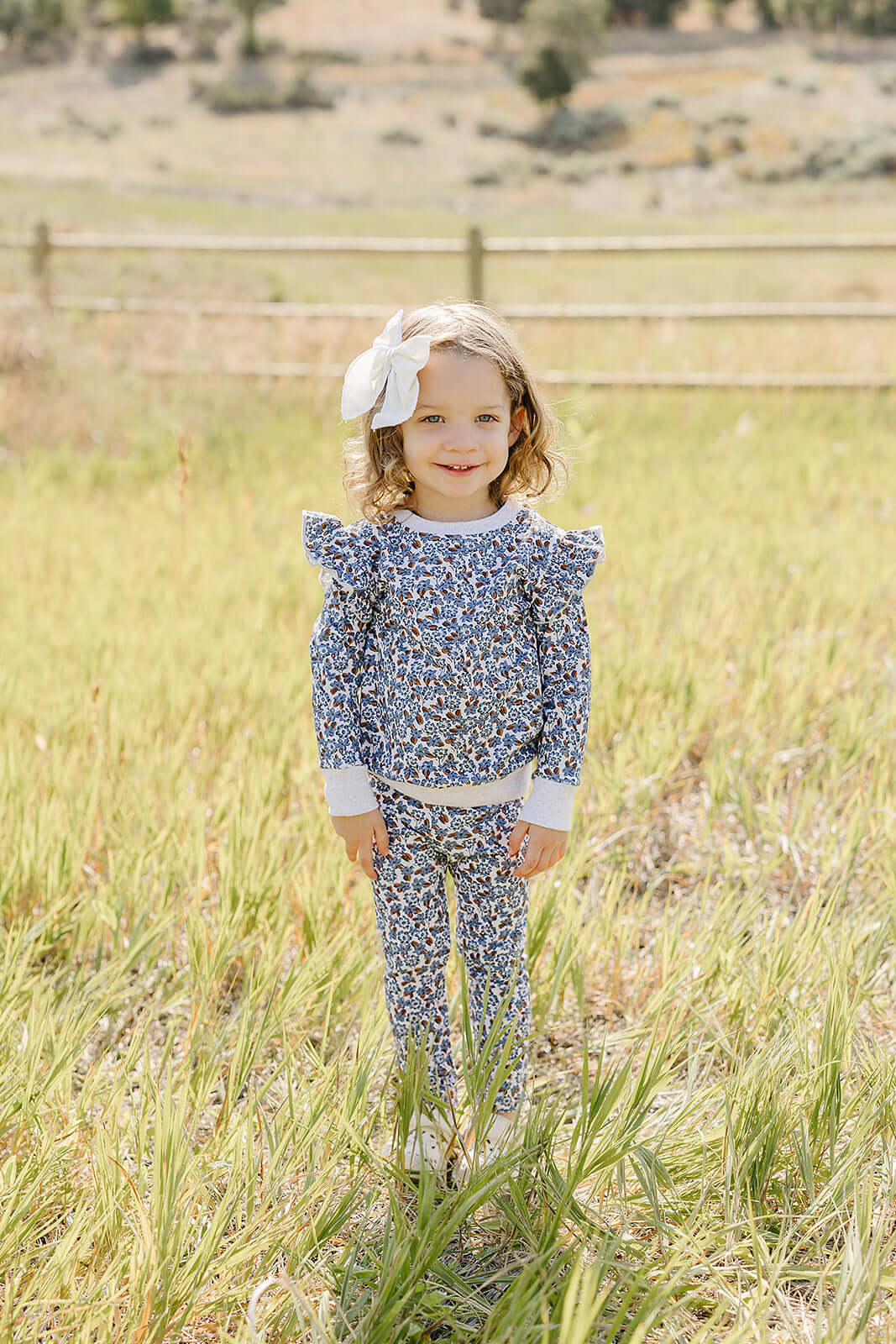 Little girl in a Blue Meadow Ruffle French Terry Set standing in a grassy field, showing off the charming floral design.