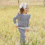 Cheerful child wearing Blue Meadow Ruffle French Terry Set, playing in a sunny field with a big bow in her hair.