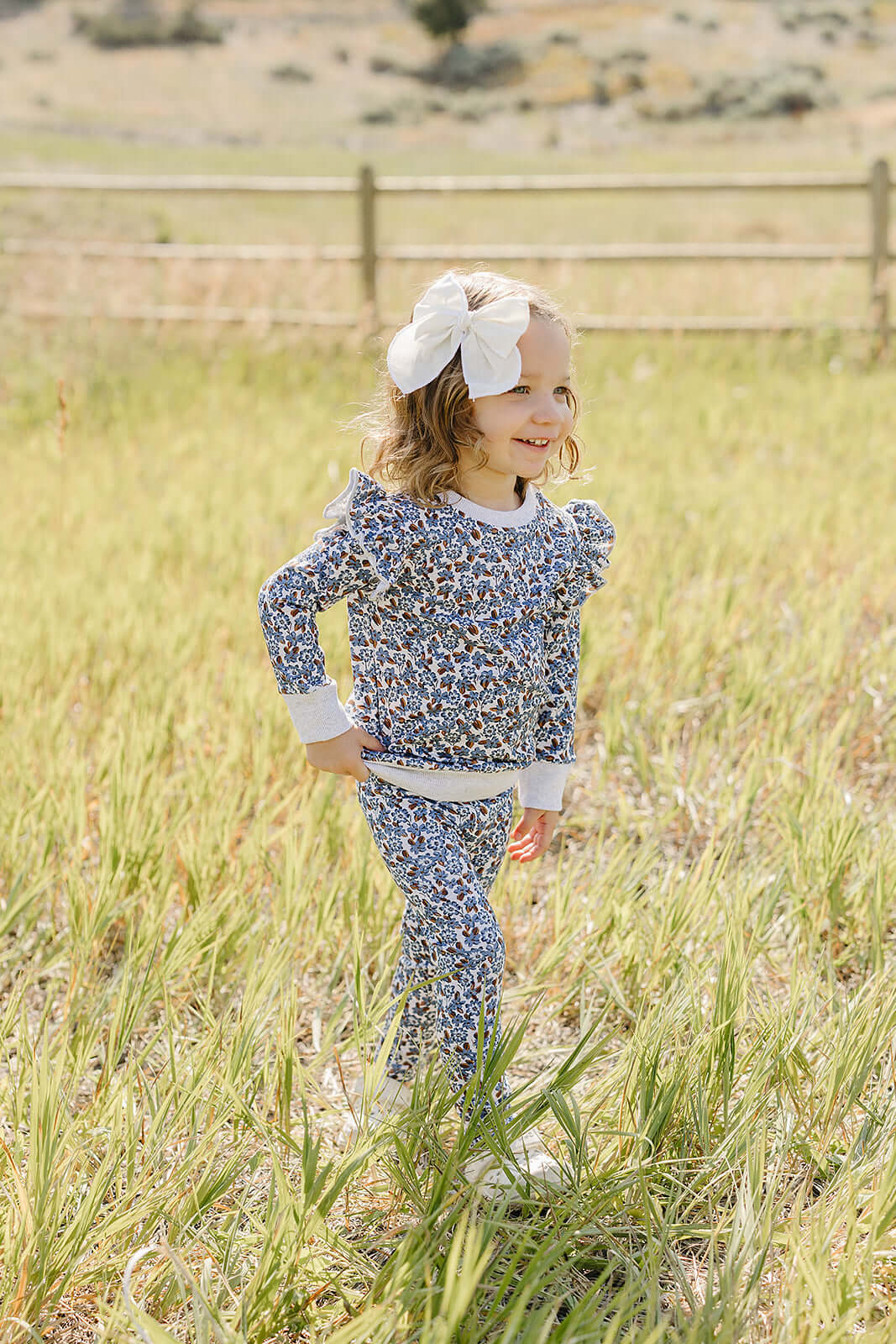 Cheerful child wearing Blue Meadow Ruffle French Terry Set, playing in a sunny field with a big bow in her hair.