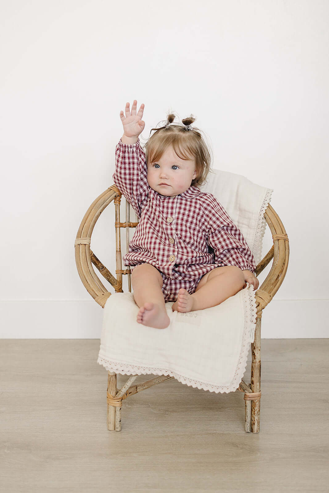 Cute baby wearing Red Gingham Peter Pan Collar Bloomer Set, sitting in a chair and raising a hand.