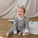 Adorable baby in Blue Meadow Ruffle French Terry Set, smiling and sitting comfortably on a woven mat.