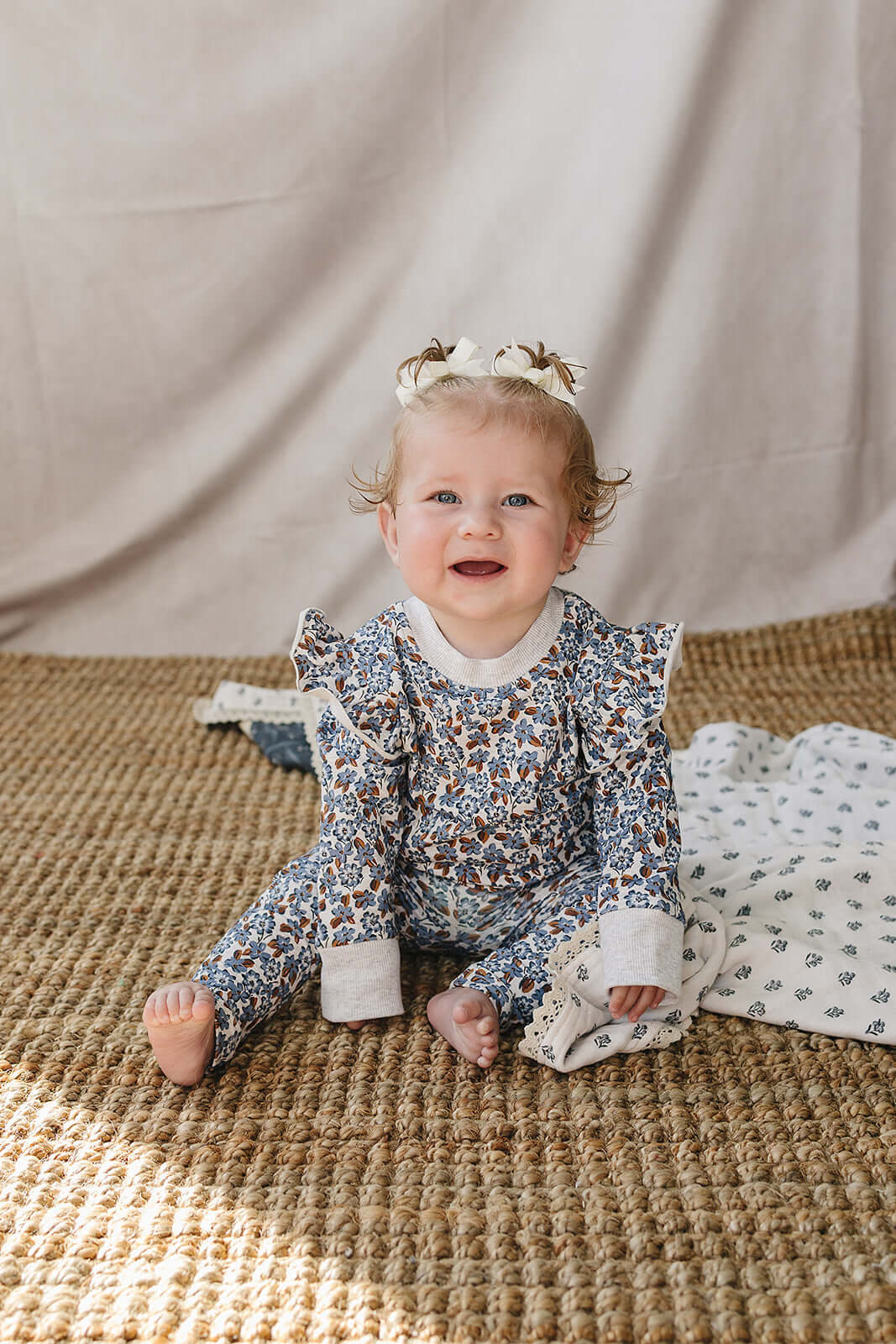 Adorable baby in Blue Meadow Ruffle French Terry Set, smiling and sitting comfortably on a woven mat.