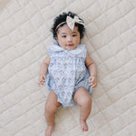 Blue Meadow Bubble Romper worn by a baby girl with curly hair on a quilted blanket.