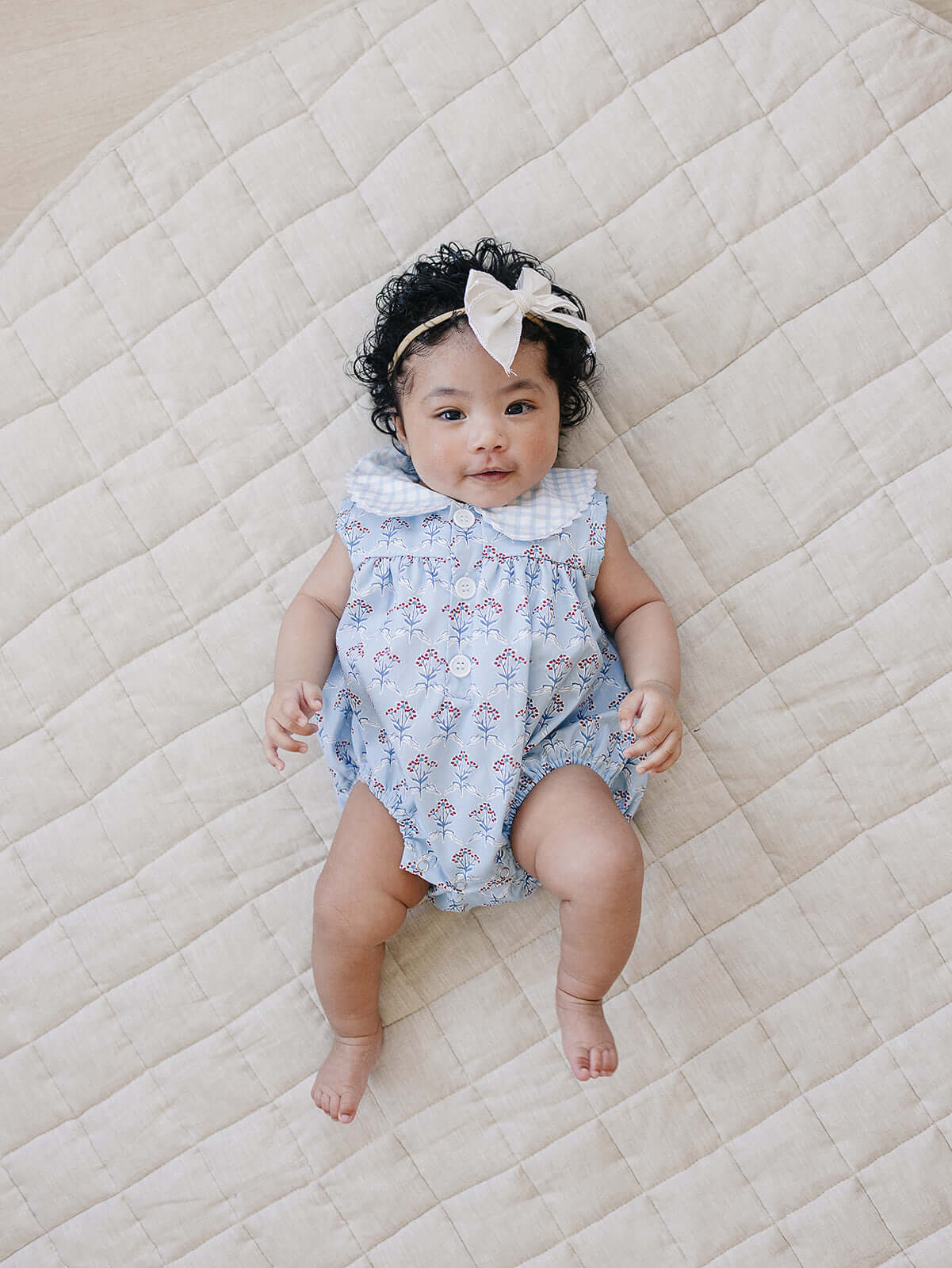 Blue Meadow Bubble Romper worn by a baby girl with curly hair on a quilted blanket.