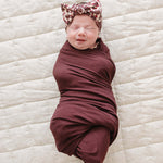 A smiling baby wrapped in a Deep Violet Ribbed Bamboo Stretch Swaddle with a floral headband, cozy and adorable.