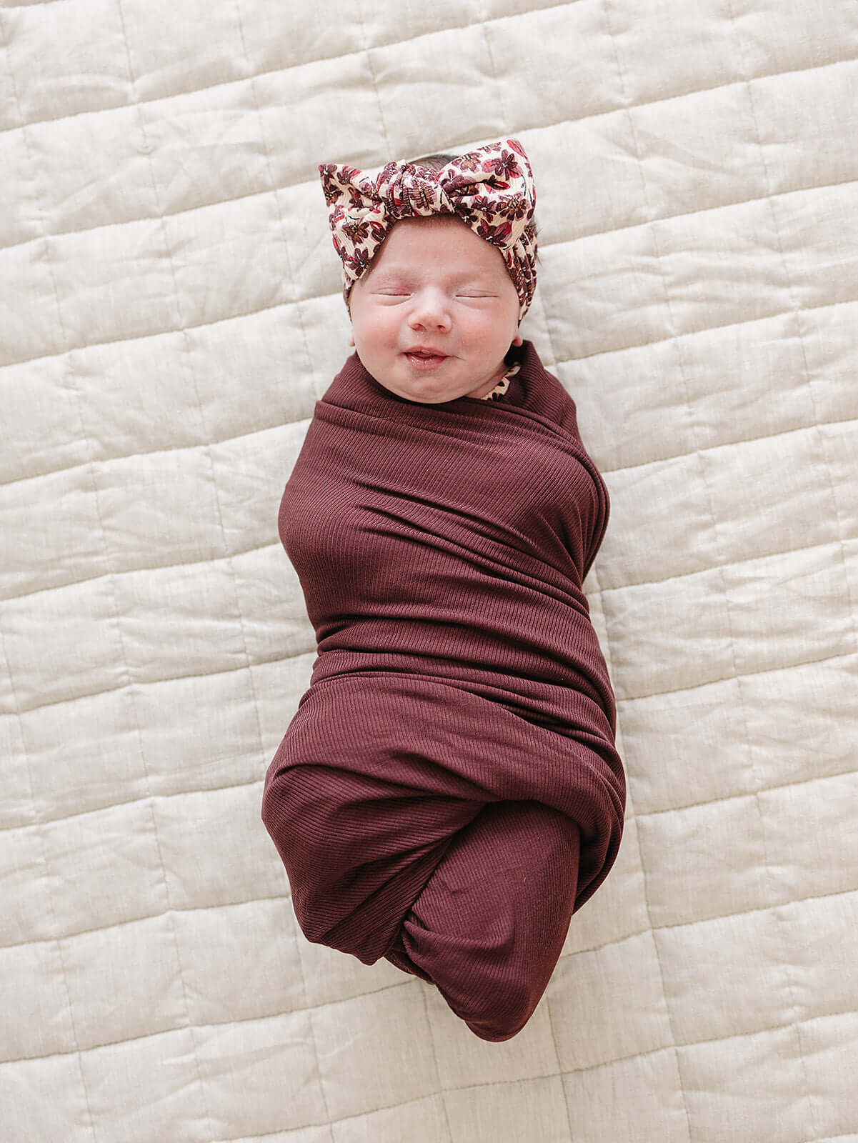 A smiling baby wrapped in a Deep Violet Ribbed Bamboo Stretch Swaddle with a floral headband, cozy and adorable.
