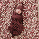 Deep Violet Ribbed Bamboo Newborn Knot Hat on a swaddled baby against a floral background.