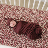 A newborn swaddled in a Deep Violet Ribbed Bamboo Newborn Knot Hat on a floral crib sheet.