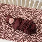 Cozy baby swaddled in Deep Violet Ribbed Bamboo Stretch Swaddle with matching hat on floral crib sheet.