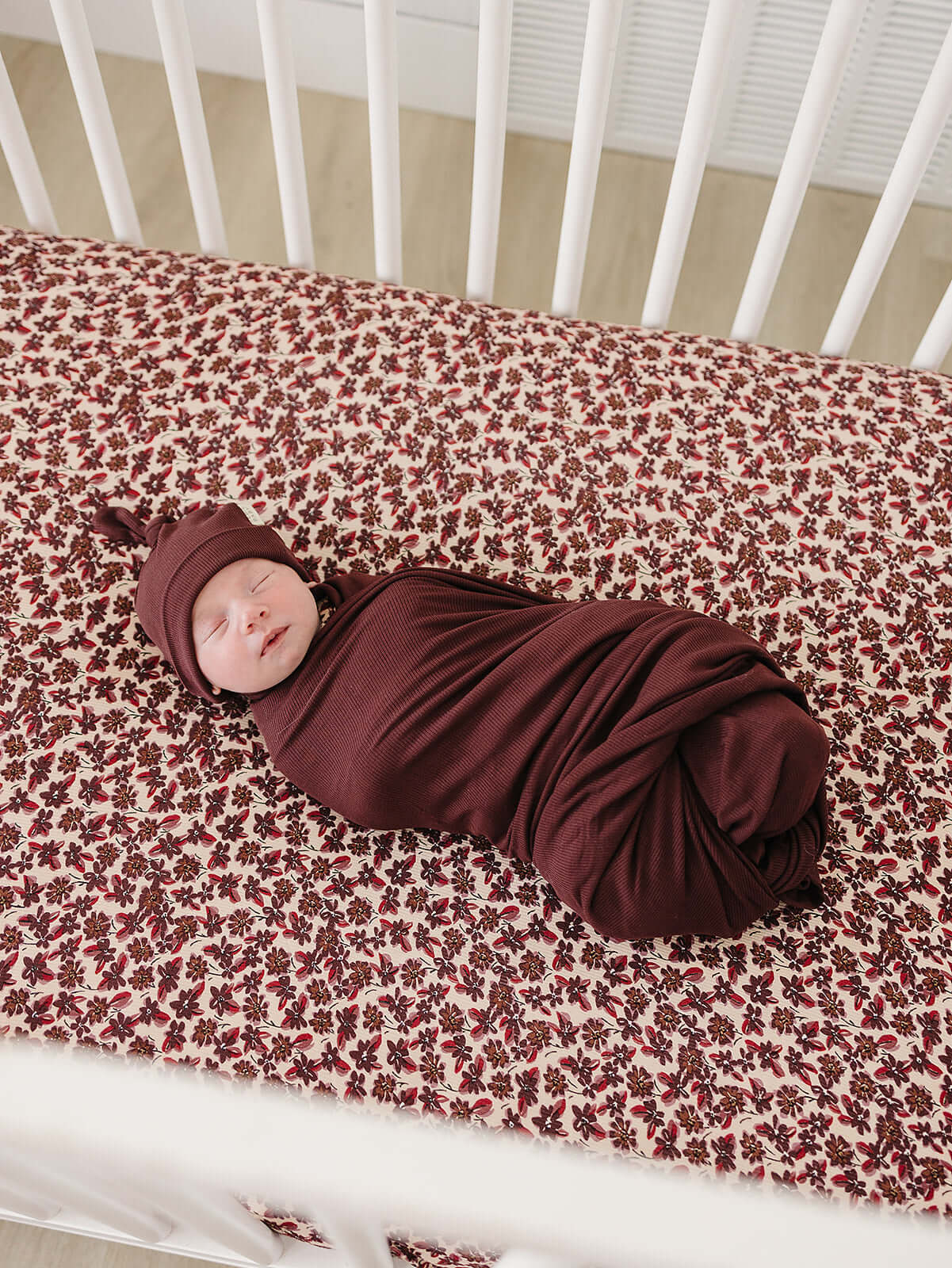 Cozy baby swaddled in Deep Violet Ribbed Bamboo Stretch Swaddle with matching hat on floral crib sheet.