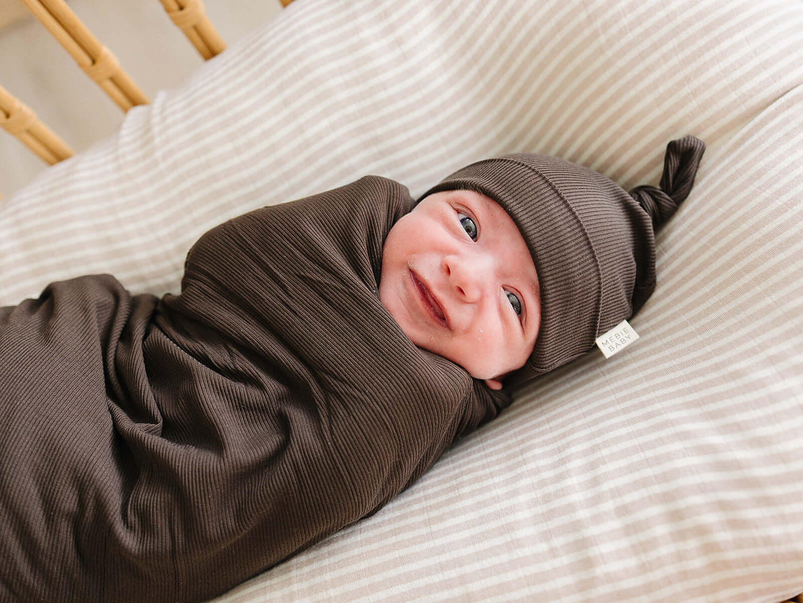 Brown Ribbed Bamboo Newborn Knot Hat on a cozy newborn, swaddled and smiling.