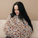Woman smiling while using Desert Floral Ribbed Bamboo Multi-Use Cover for nursing on a couch.