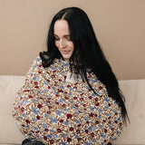 Woman smiling while using Desert Floral Ribbed Bamboo Multi-Use Cover for nursing on a couch.