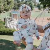 Baby wearing Americana | Ribbed Bamboo Zip Pajamas with a bow headband outdoors, showcasing fun American flag design.
