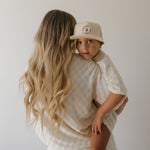 Woman holding a child wearing a beige cap with a logo against a plain background
