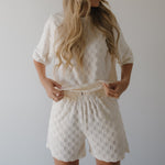 Person wearing a white textured outfit with shorts against a plain background