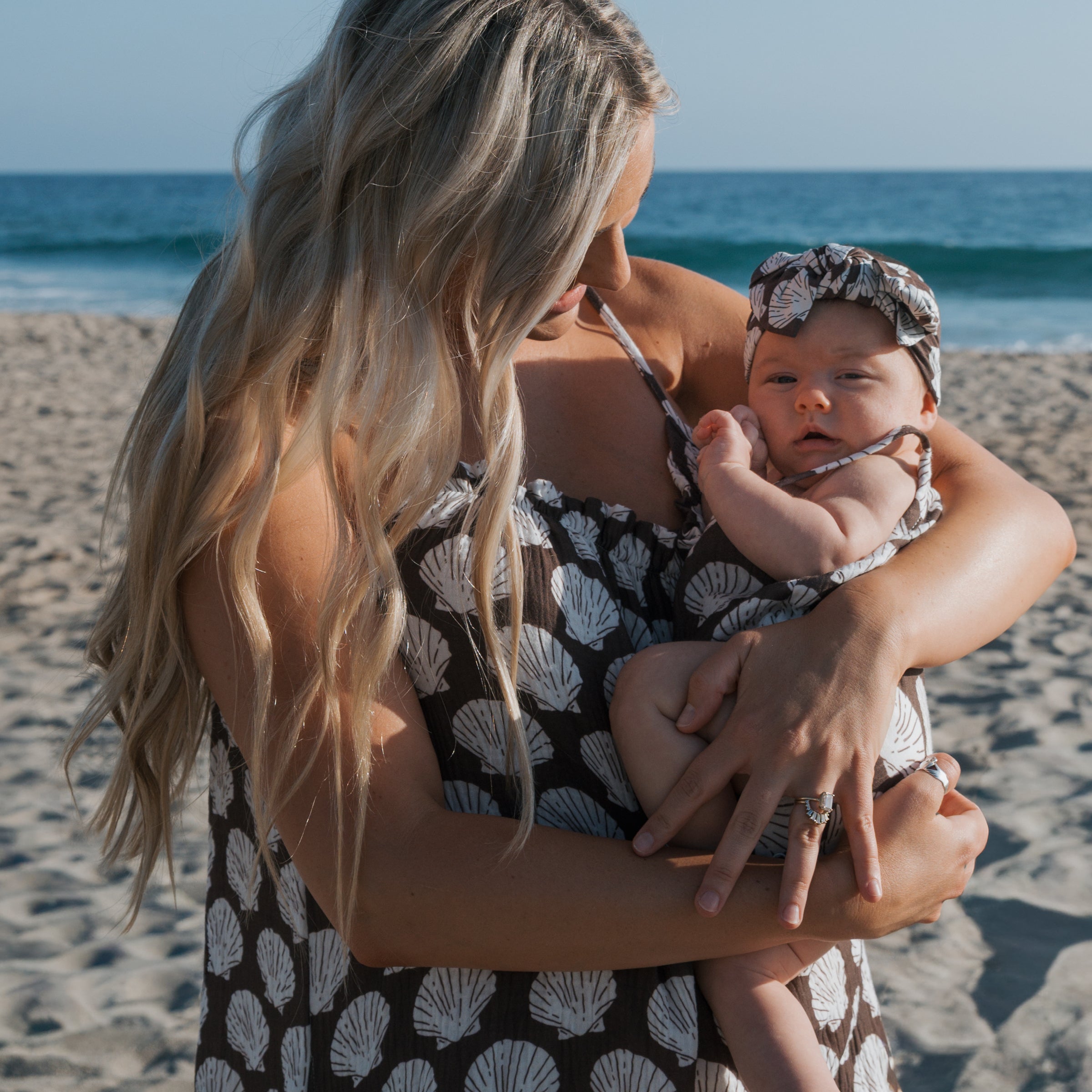 Mom holding baby in matching seashell muslin tank romper at the beach, perfect for warm days.