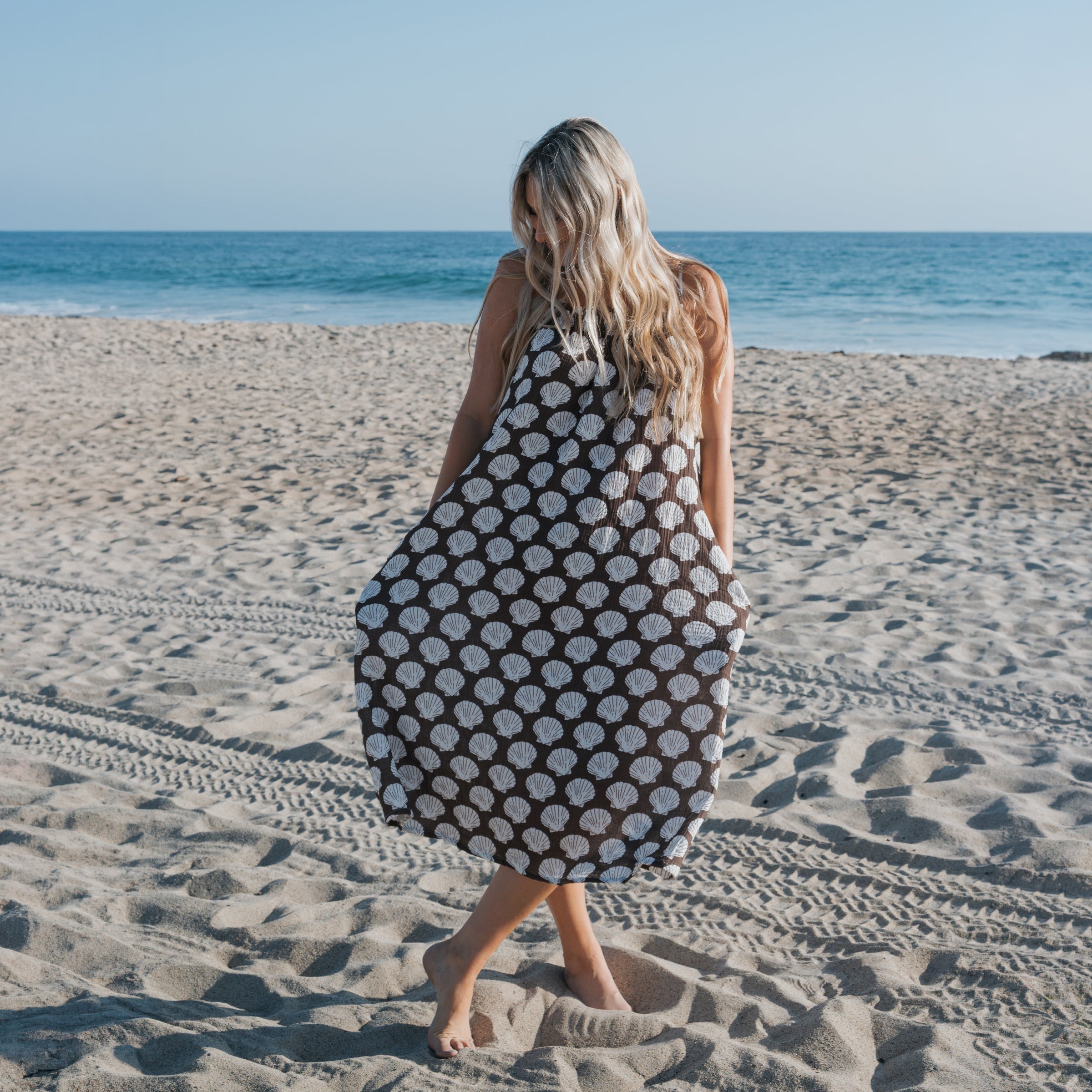 Woman wearing a Women's Muslin Sleeveless Dress with seashell print standing on the beach by the ocean.