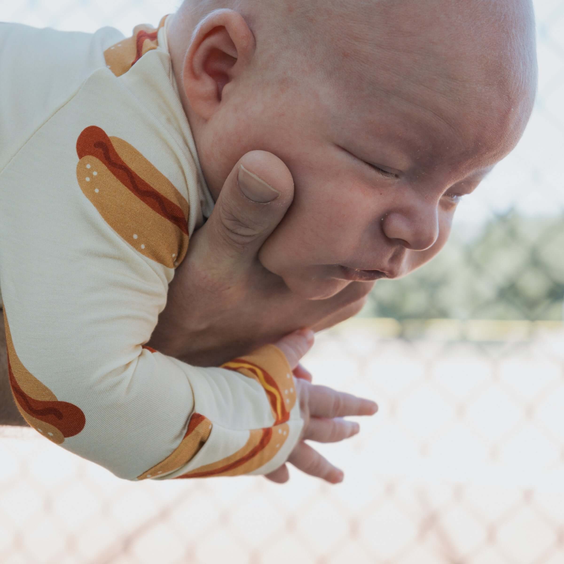 Baby in Hot Dogs | Bamboo Zip Pajamas, held by an adult, in a playful outdoor setting.