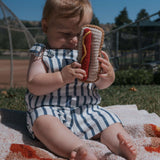 A baby sitting on a blanket holding a Knit Rattle | Hot Dog toy, enjoying summer fun outdoors.
