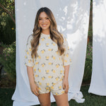 Woman wearing a lemon-patterned outfit standing in front of white curtains with greenery in the background