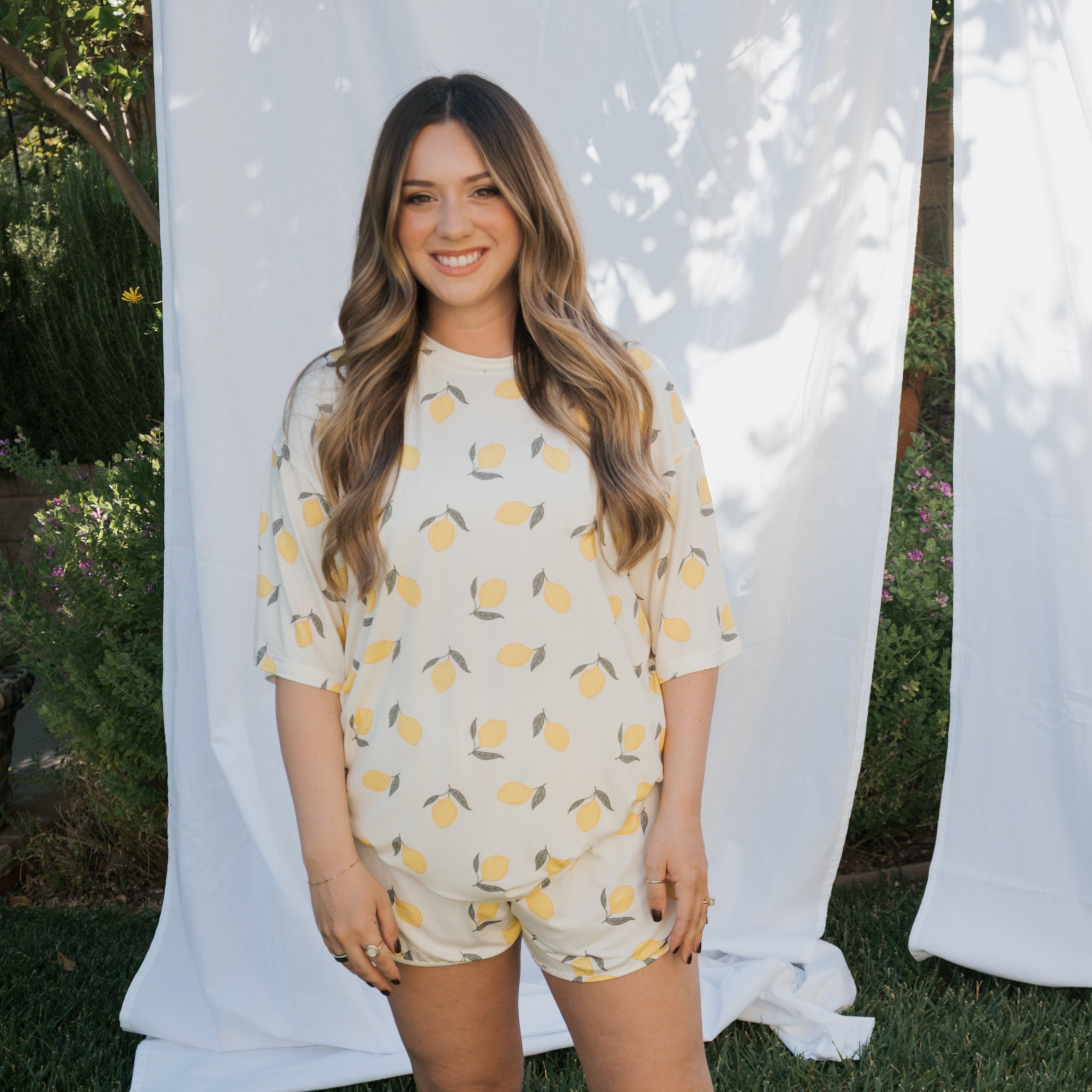 Woman wearing a lemon-patterned outfit standing in front of white curtains with greenery in the background