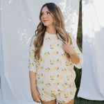 Woman wearing a yellow and white patterned outfit standing outdoors with white curtains in the background.
