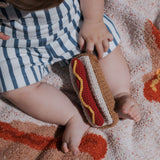 A baby happily shaking a Knit Rattle | Hot Dog on a cozy blanket, adding cheer to summer fun!