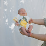 Baby wearing a onesie with a yellow flower design, held by an adult against a white background.