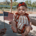 A baby wearing a Picnic Pinstripe | Knit Ruffle Romper, holding a small American flag, enjoying a sunny day.