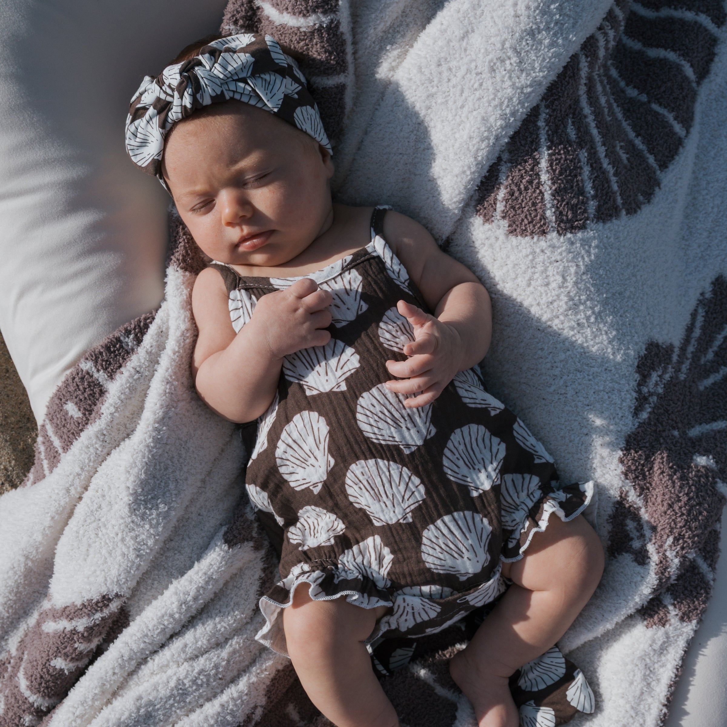 Baby wearing a seashell patterned muslin tank romper and matching headband, relaxing on a soft blanket.