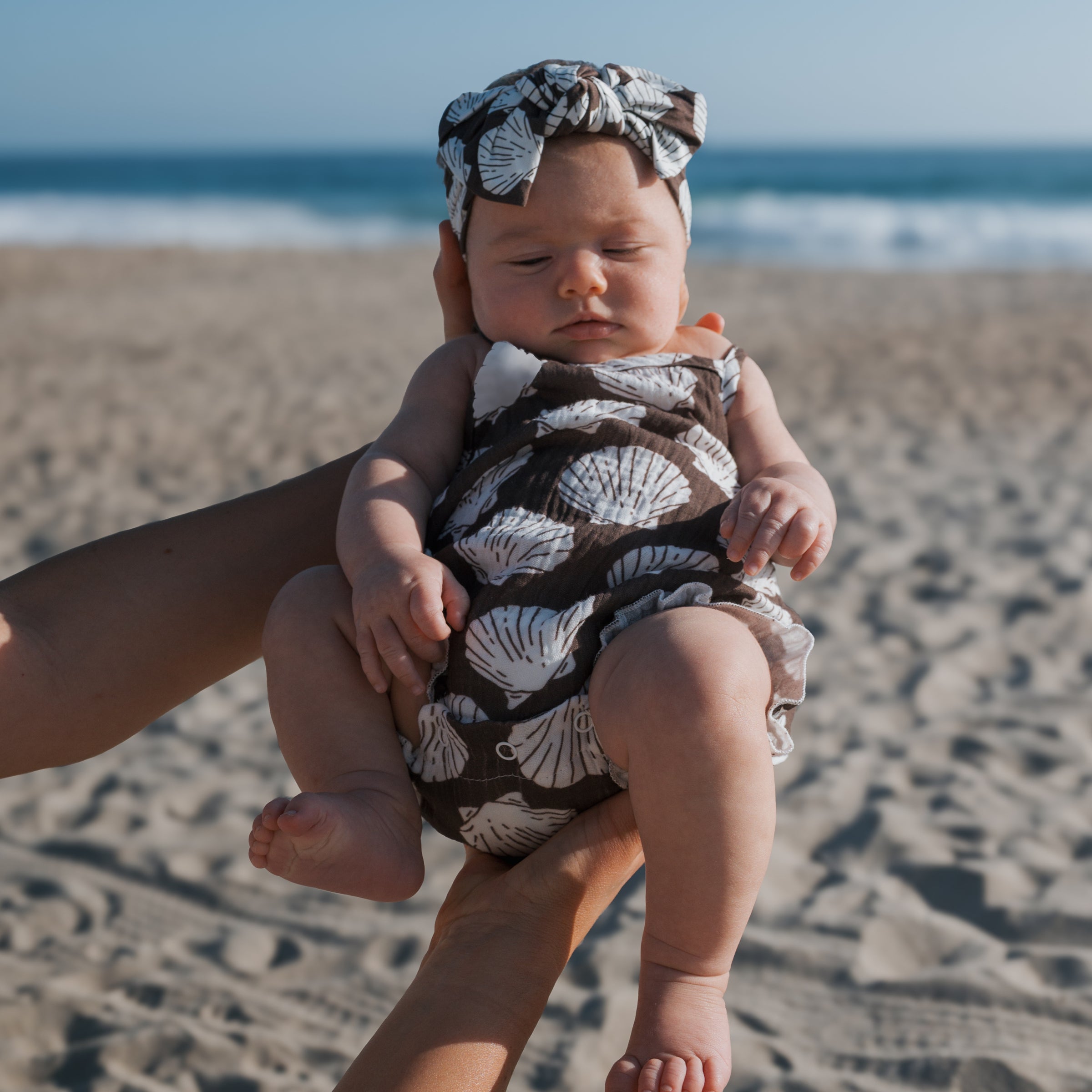 Baby wearing seashell print muslin tank romper at the beach, perfect for warm days and matching styles.