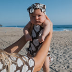 Baby wearing a muslin tank romper with seashell pattern and matching bow, held by an adult on a beach.
