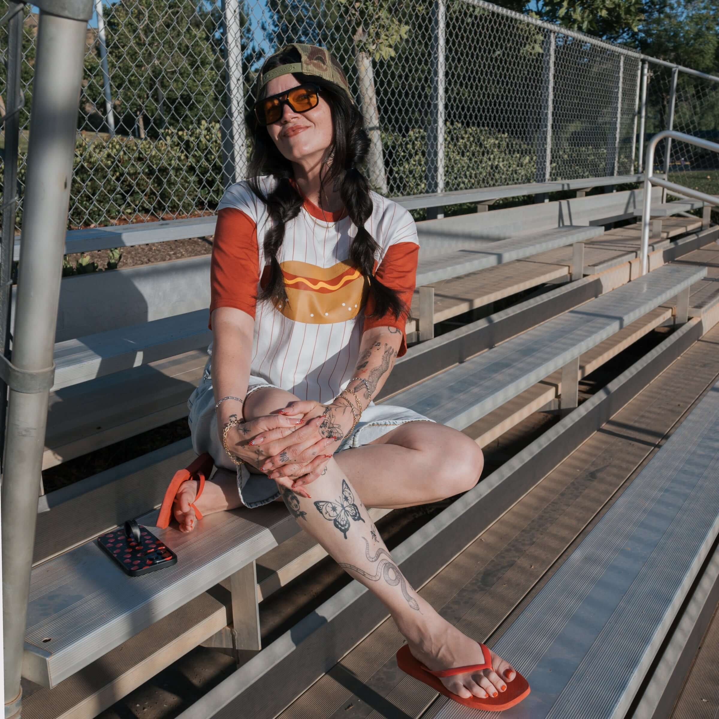 A model wearing a Hot Dogs | Adult Cotton Tee sitting on bleachers, enjoying a sunny day with sunglasses.