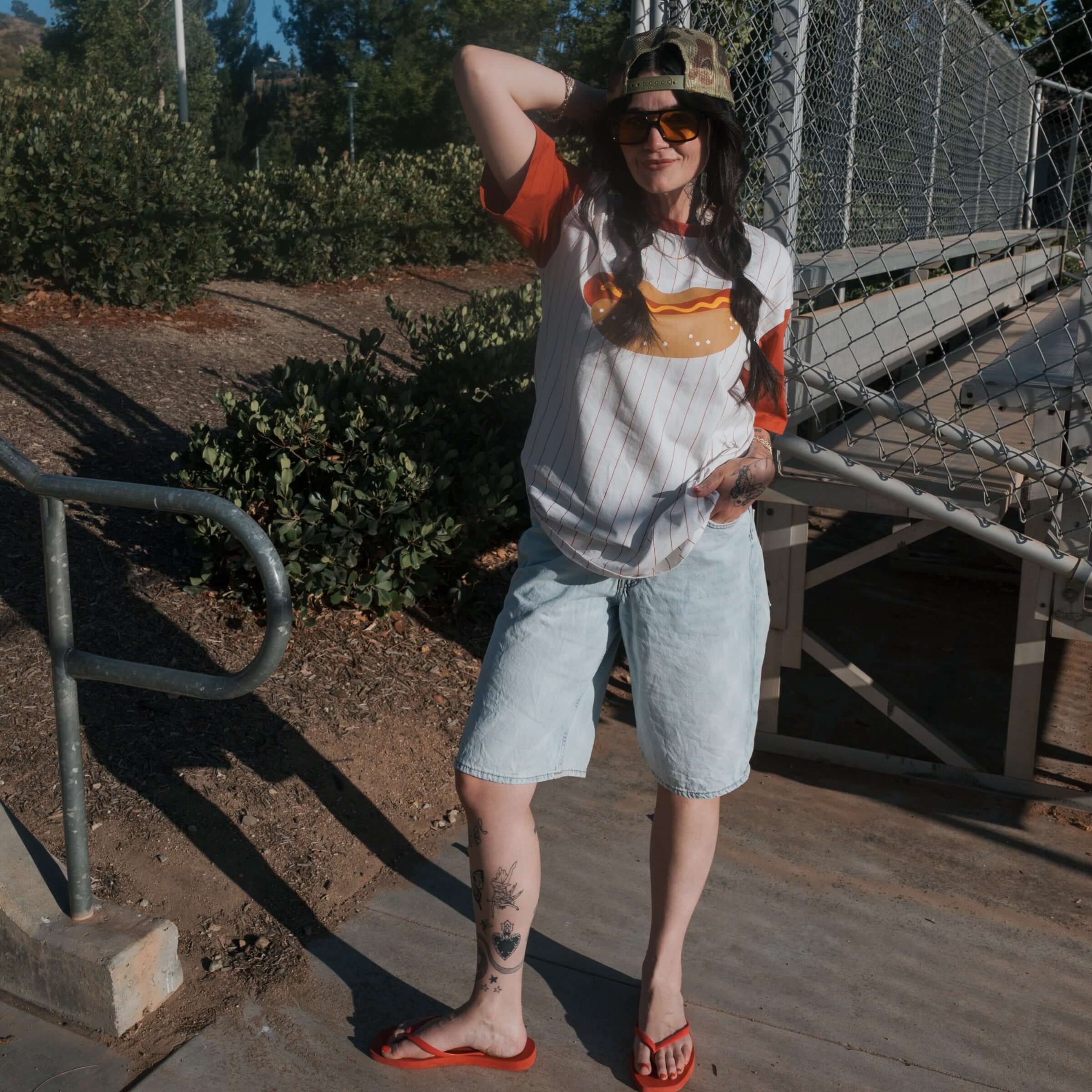 A model showcasing the Hot Dogs | Adult Cotton Tee paired with denim shorts and flip-flops in a casual outdoor setting.