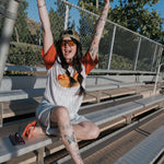 A joyfully posed woman wearing a Hot Dogs | Adult Cotton Tee, sitting on bleachers with arms raised in excitement.