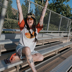 A joyfully posed woman wearing a Hot Dogs | Adult Cotton Tee, sitting on bleachers with arms raised in excitement.