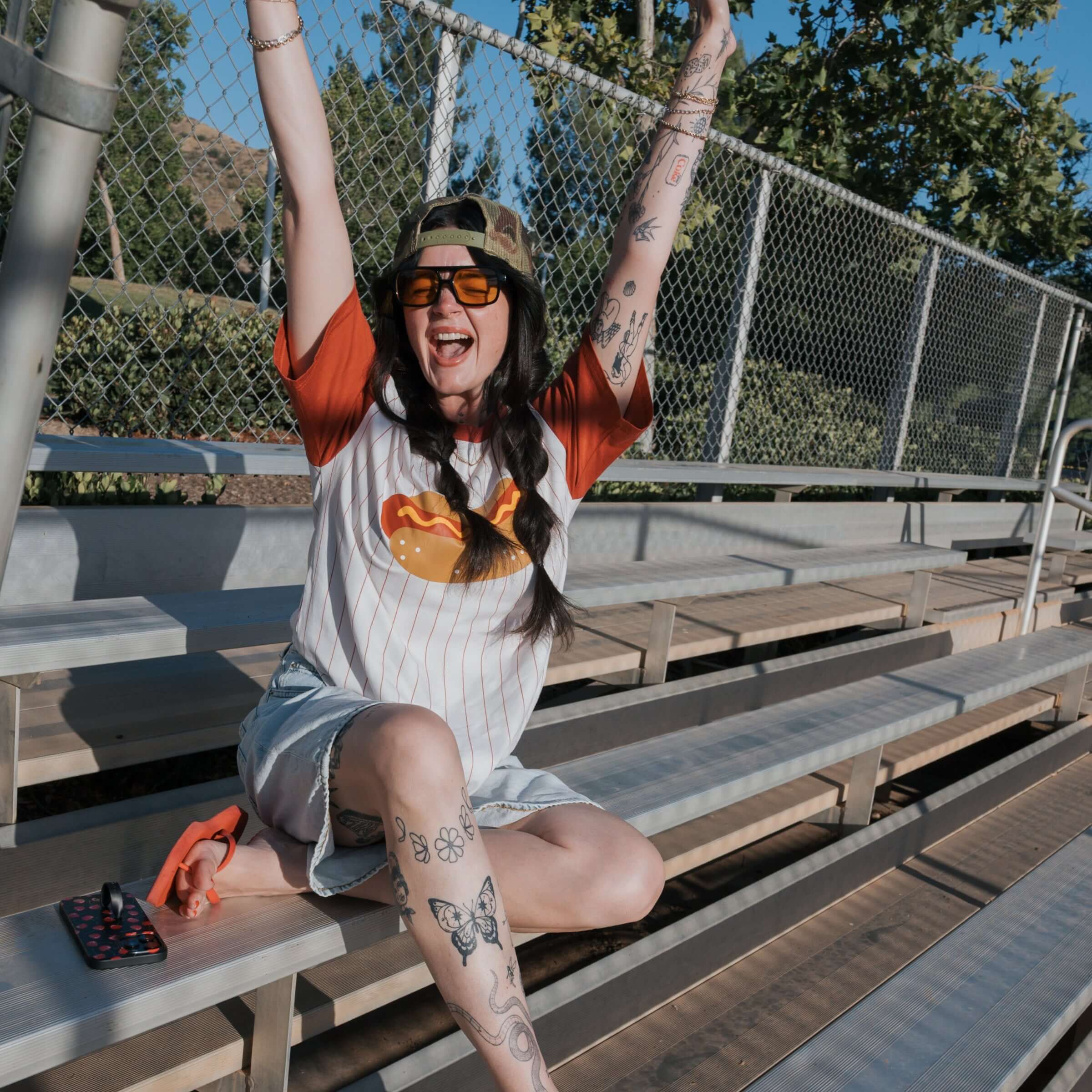 A joyfully posed woman wearing a Hot Dogs | Adult Cotton Tee, sitting on bleachers with arms raised in excitement.