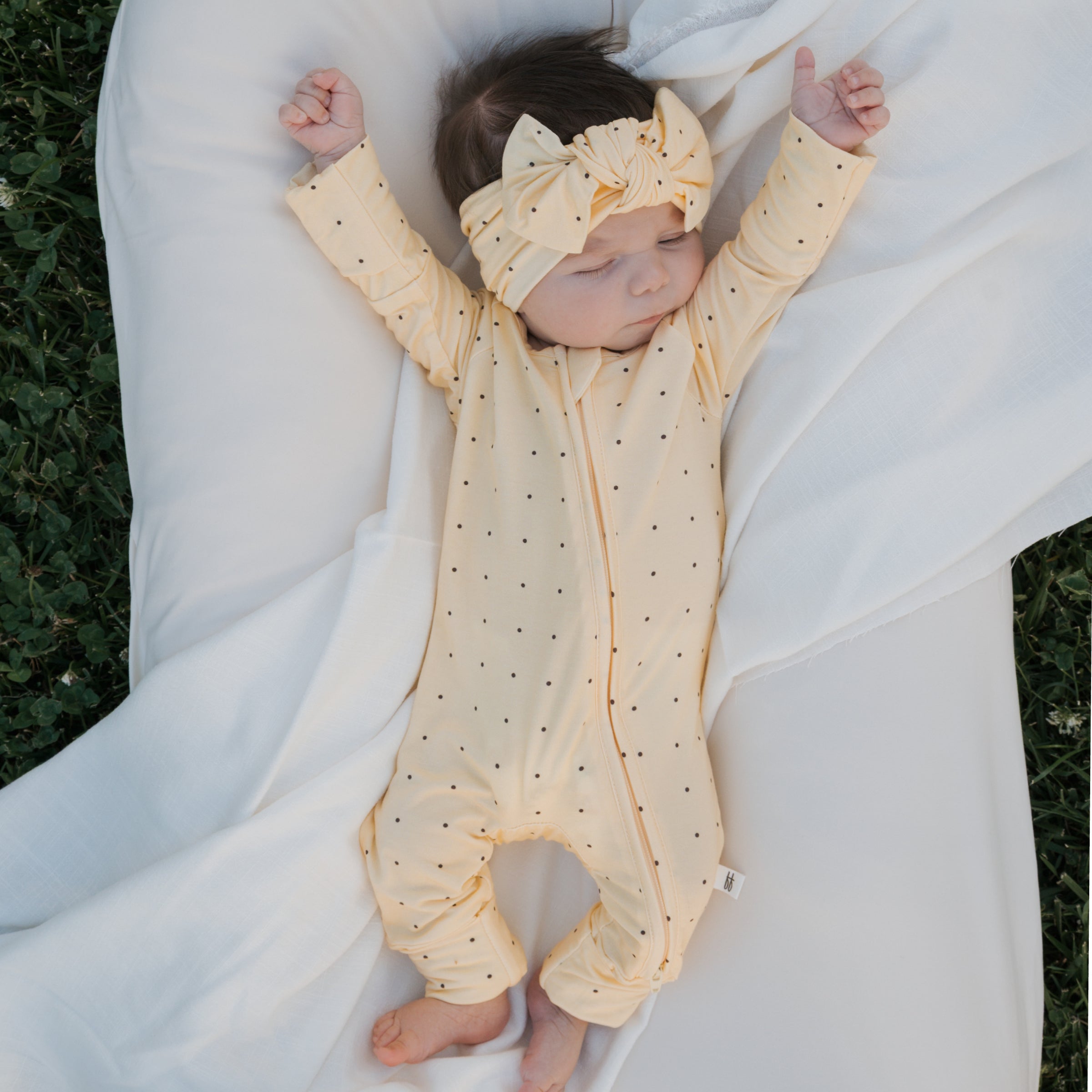 Baby wearing Veda Bamboo Zip Pajamas in yellow with black polka dots, relaxing on a white blanket outdoors.