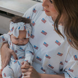 A mother holding her baby wearing an Americana | Ribbed Bamboo Head Wrap, showcasing a cute bow and American flag print.